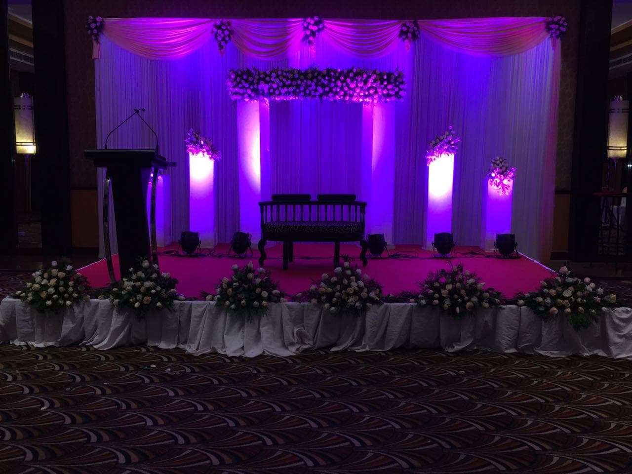 Purple-lit wedding stage with white drapes and roses in a ballroom